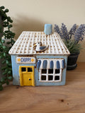Decorative house-shaped tea light with a yellow door and blue accents on a wooden surface.